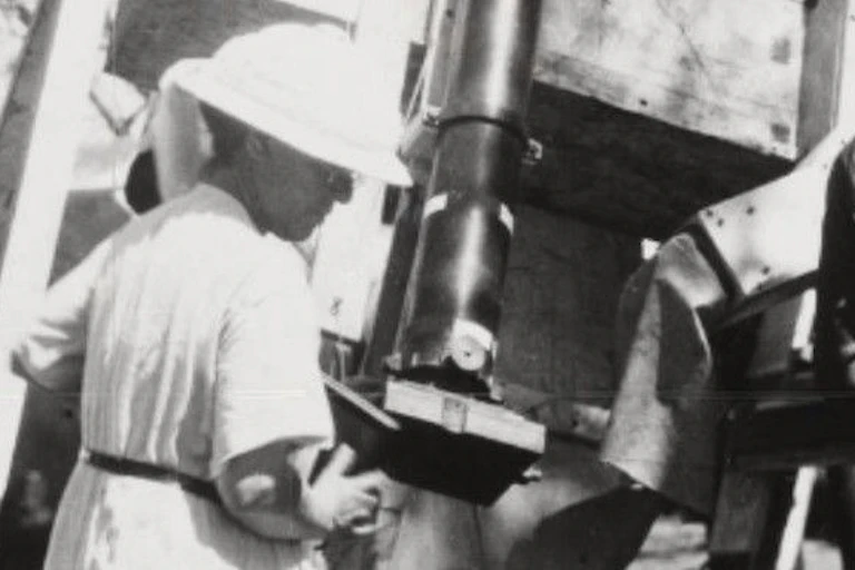 A woman in a white dress and hat operating a telescope on a wooden frame
