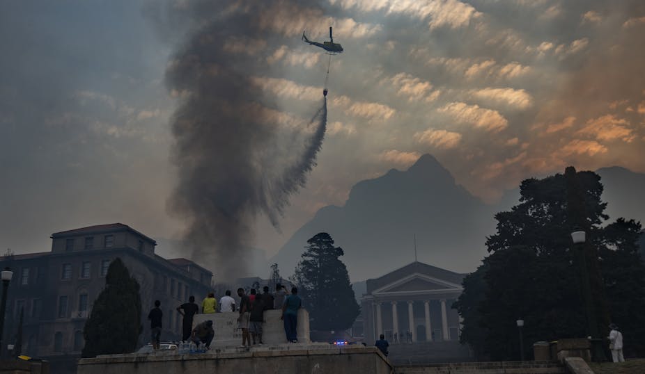 Smoke rises from buildings and a helicopter releases water. Mountain in the background, trees and people in the foreground.