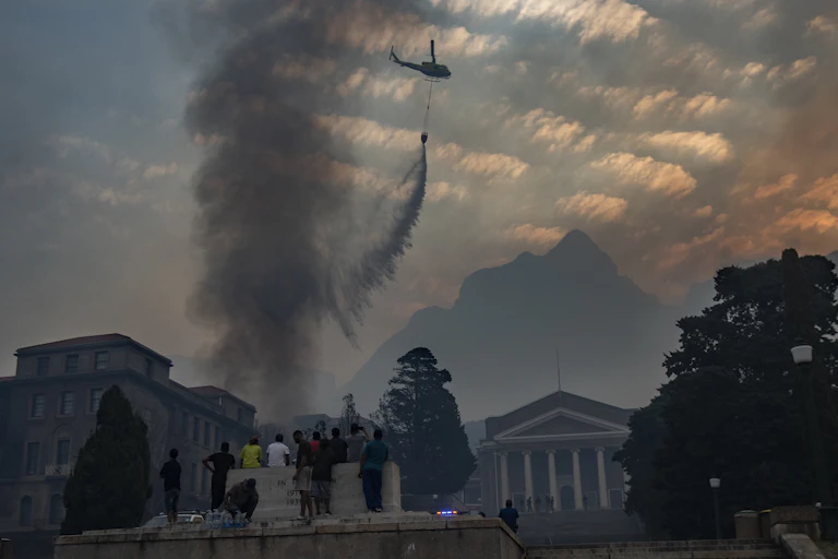 Smoke rises from buildings and a helicopter releases water. Mountain in the background, trees and people in the foreground.