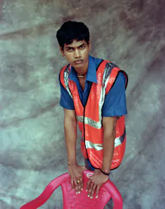 A man leans on a red plastic chair, wearing a high vis jacket decorated in sequins.