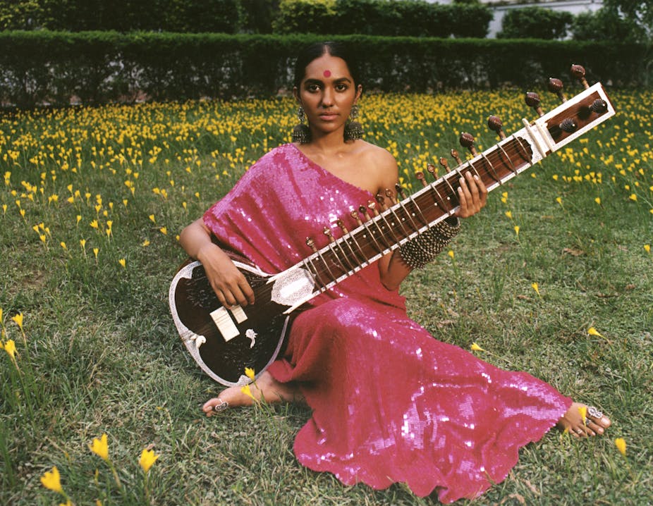 A woman plays a sitar in a field, wearing a bright pink sequinned sari
