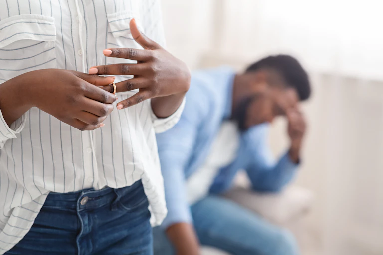 woman removing wedding ring from finger, her despairing husband on background