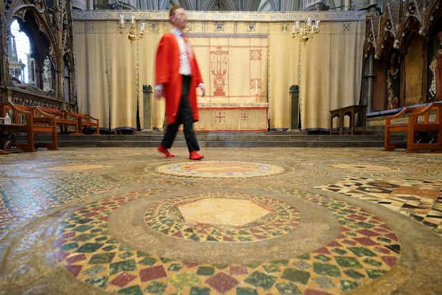 An abbey marshall in a red coat and red socks walks across a mosaic floor in Westminster Abbey.
