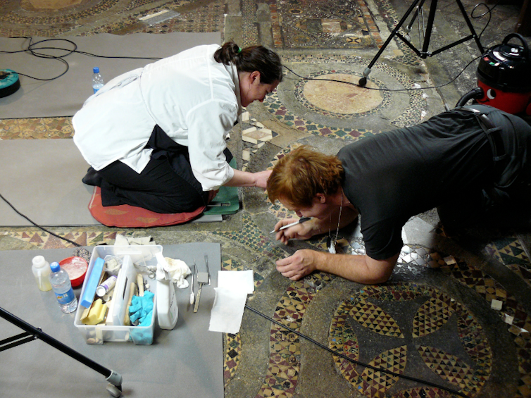 Two people with tools kneel on a mosaic floor.
