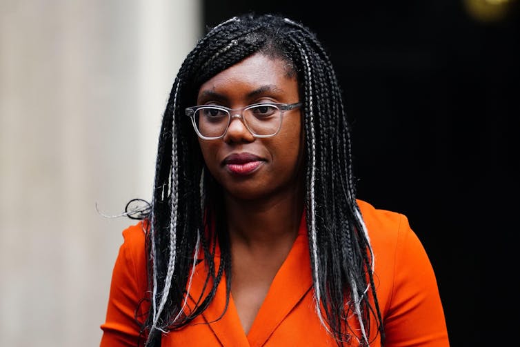 UK business and trade secretary Kemi Badenoch, organge jacket, glasses, with a black door in the background.