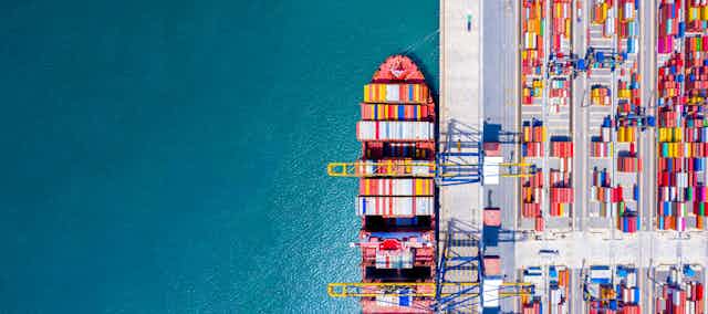 Aerial view of a ship docked at a port with shipping containers on one side and blue sea on the other.