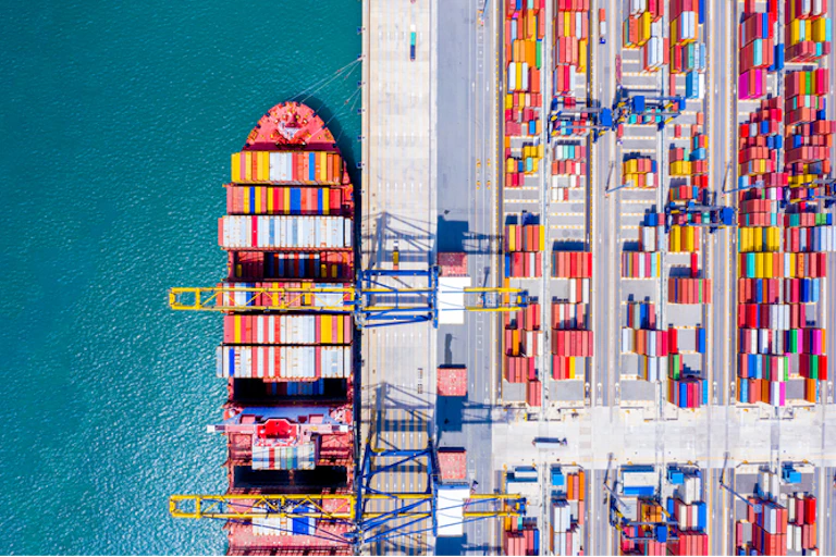 Aerial view of a ship docked at a port with shipping containers on one side and blue sea on the other.