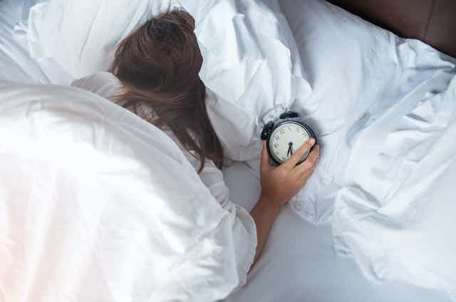 Mujer acostada en la cama boca abajo con el despertador en la mano.