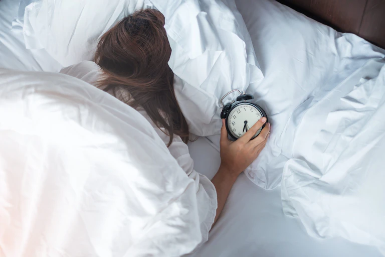 Mujer acostada en la cama boca abajo con el despertador en la mano.
