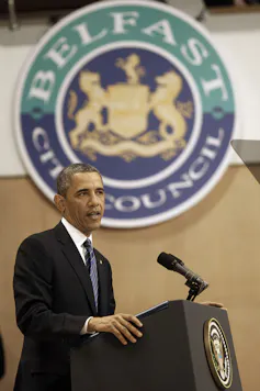 Side view of Barack Obama standing at a podium, with a Belfast City Council crest on the wall behind him.