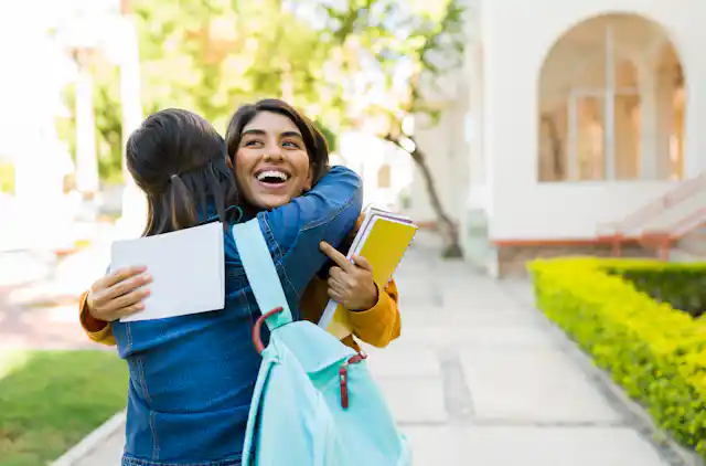 A person holding notebooks and a letter smiles and hugs a person carrying a backpack.