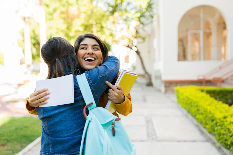A person holding notebooks and a letter smiles and hugs a person carrying a backpack.