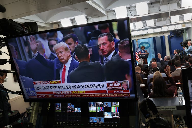 A TV screen in a press room shows a white man waving, with the words'Now: Trump is being processed against arraignment.'