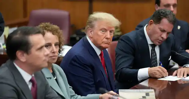 A rotund blond man in a blue suit and red tie sits in a courthouse with his legal team.