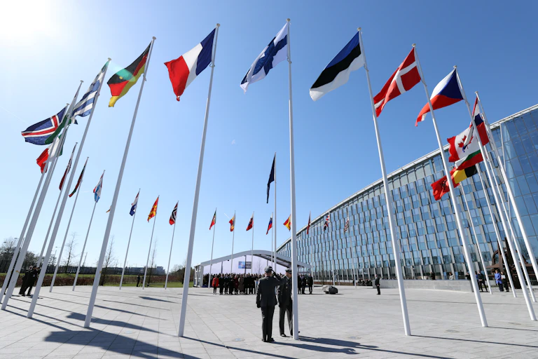 The Finnish flag stands among those of other countries outside a glass building. Two men salute in uniform underneath.