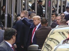 A white man with white hair and a dark suit waves, as he is surrounded by men in dark suits.