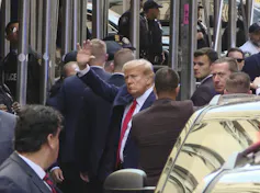 A white man with white hair and a dark suit waves, as he is surrounded by men in dark suits.
