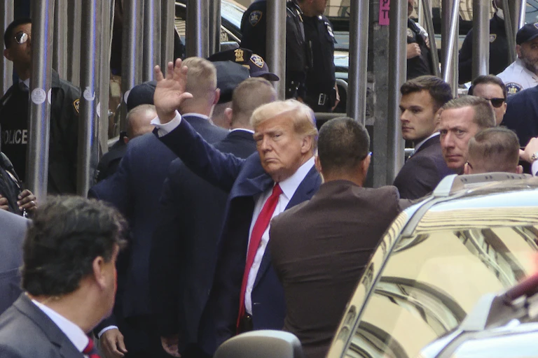 A white man with white hair and a dark suit waves, as he is surrounded by men in dark suits.