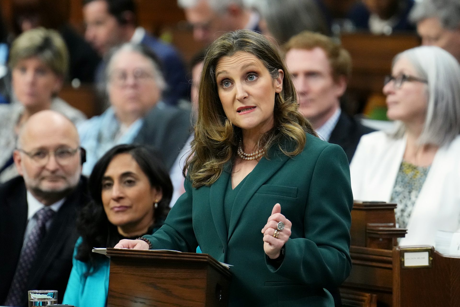 A white middle-aged woman in a green suit gestured while speaking from behind a podium. A crowd of people are seated behind her.