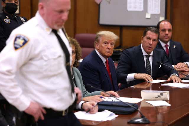 A white man with blue suit sits at a table with a solemn face, surrounded by three other people and a man standing in a white shirt with a police badge. 