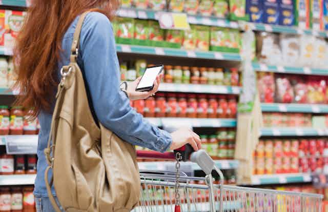 A woman, seen from slightly behind, looking at a phone screen while pushing a shopping cart down an aisle