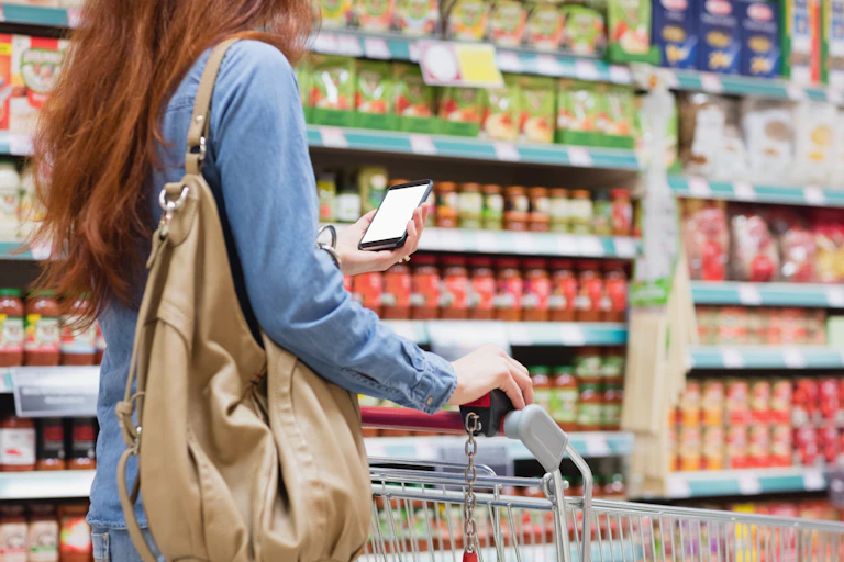 A woman, seen from slightly behind, looking at a phone screen while pushing a shopping cart down an aisle