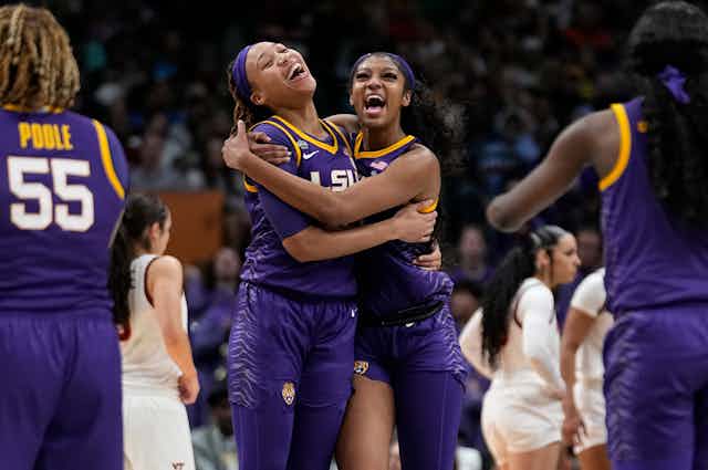 Two female basketball players, dressed in a purple uniform, embrace on the basketball court.