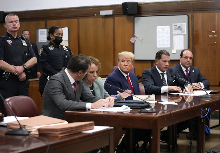 A suited man sits at a courtroom table with uniformed court officers standing behind him and attorneys sitting on his left and right.
