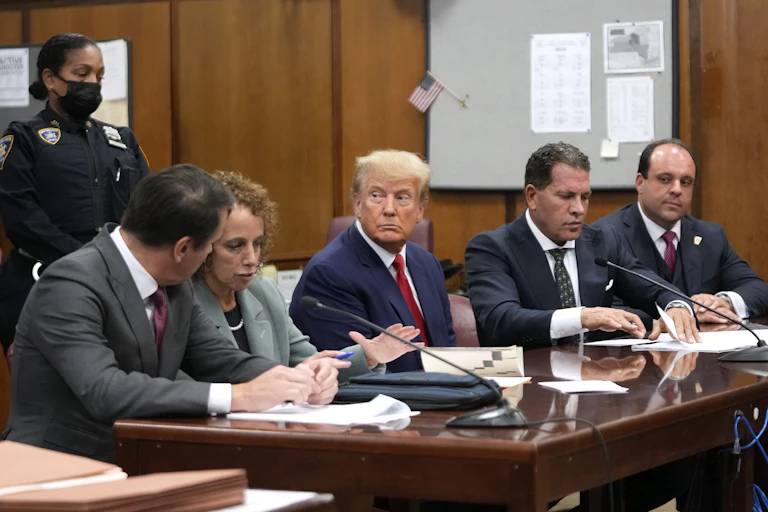 A suited man sits at a courtroom table with uniformed court officers standing behind him and attorneys sitting on his left and right.