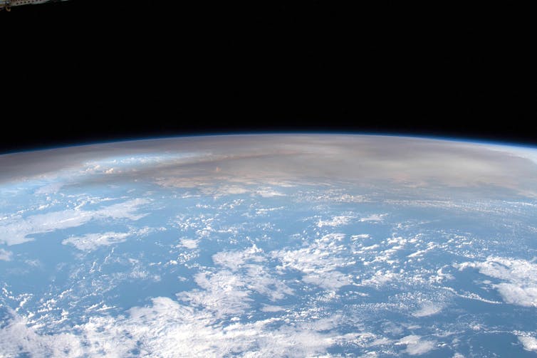 A photo from the International Space Station showing white puffy clouds over the ocean and a dark grey plume from a volcanic eruption.