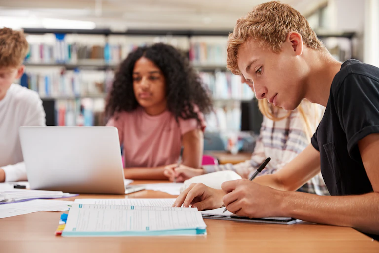 Teenagers work at a library desk with books and a laptop.