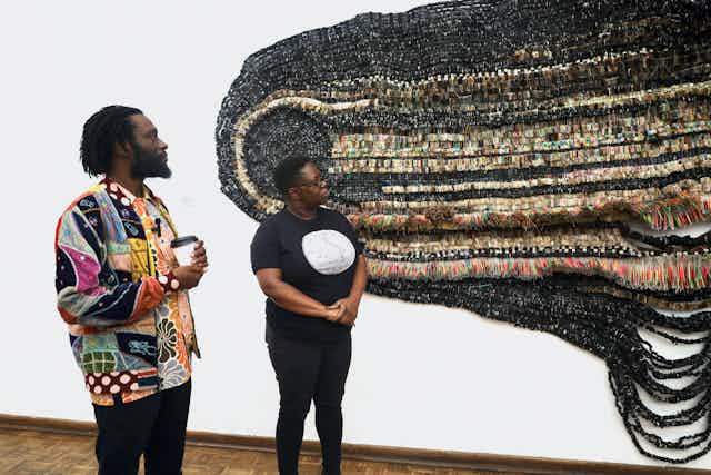 A man in a colourful jacket, with a beard and dreadlocks stands with a woman in jeans and a T-shirt looking at a large tapestry-like artwork on a wall.