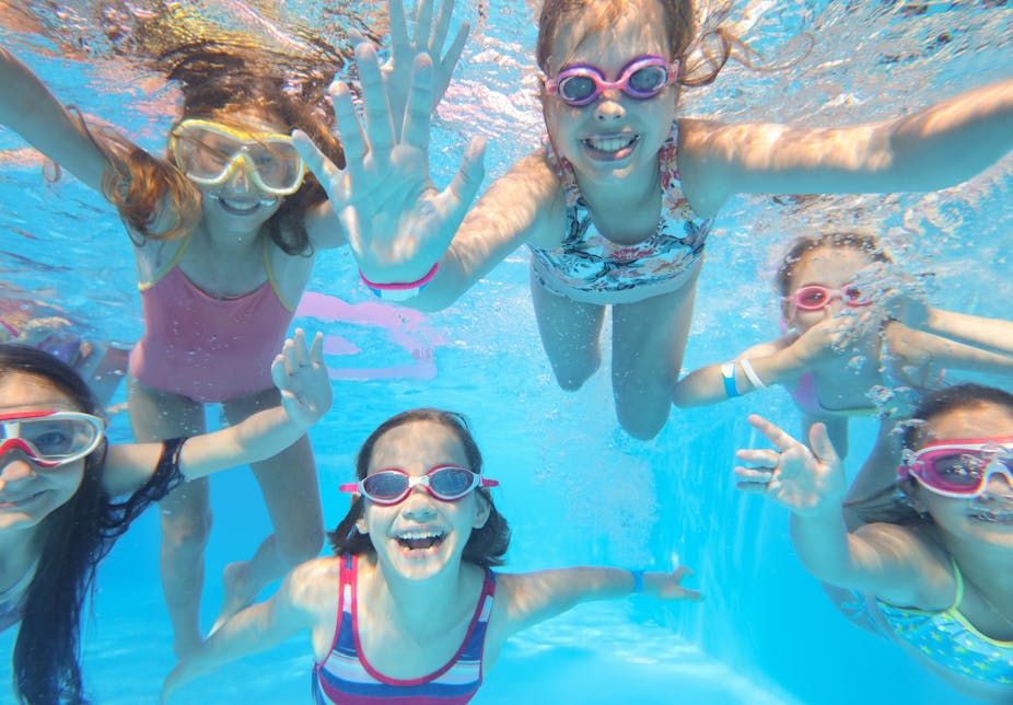 Children wearing goggles swim underneath the water of a swimming pool. They are happy and waving at the camera. 