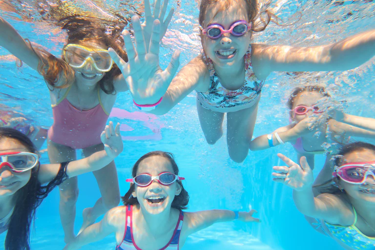 Children wearing goggles swim underneath the water of a swimming pool. They are happy and waving at the camera.