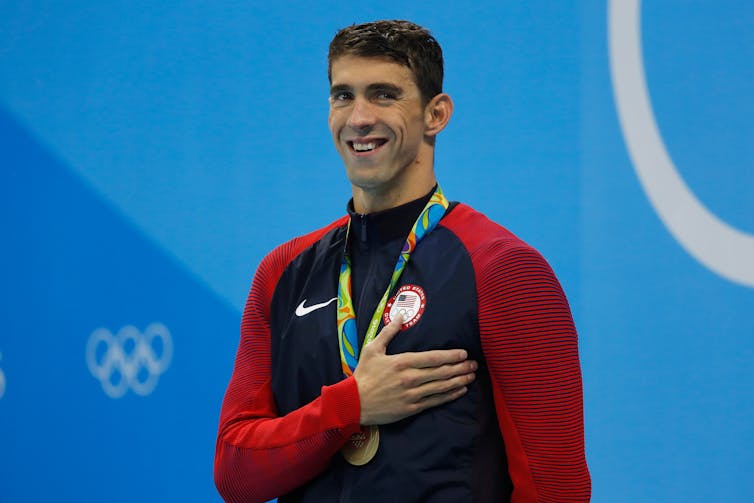 A smiling man with a gold medallion around his neck holds his right hand over his heart.