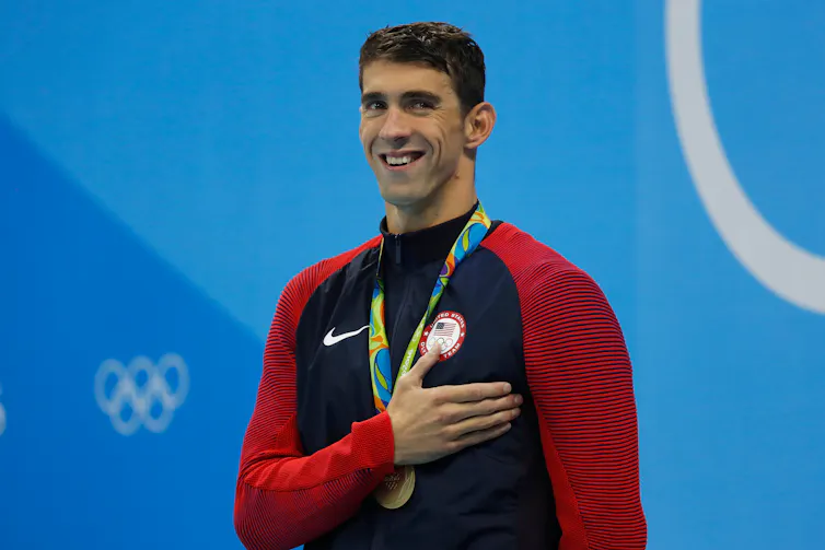 A smiling man wearing a gold medal around his neck holds his right hand over his heart.