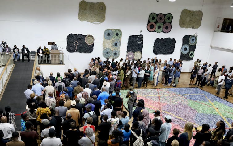 A crowd of people listens to a woman giving a speech in an art gallery, with eight large tapestry-like sculptures mounted on a wall.