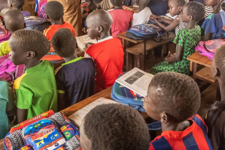 Several children sitting in a classroom with bags on long desks that sit four children, and some school books on top of the bags
