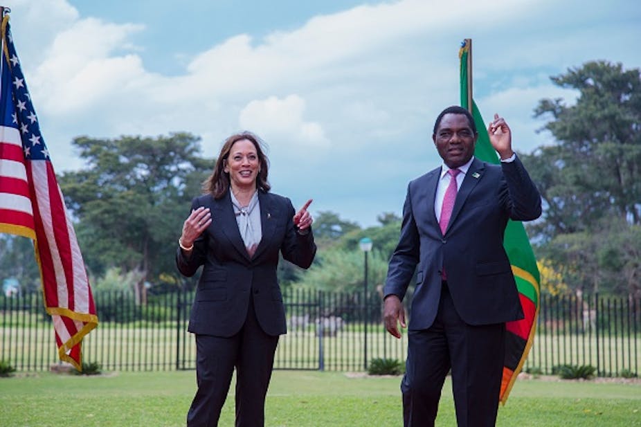 A man and woman pointing forward while standing in a field, with the Zambian and American flags behind them. 