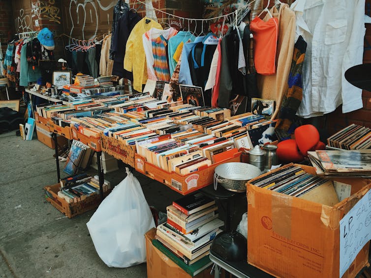 a stall at a secondhand market with books on a table, clothes on a hangline, and household objects piled up