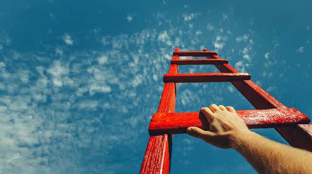 Picture taken from below of a hand on a red ladder rising to a blue sky with clouds.