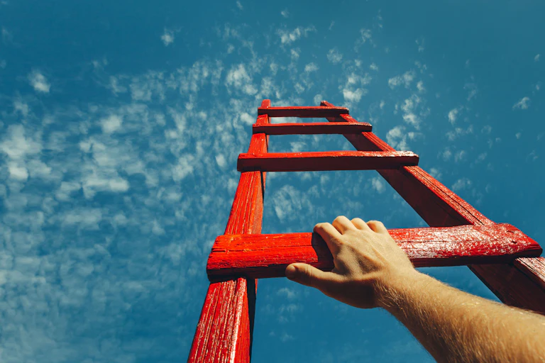Picture taken from below of a hand on a red ladder rising to a blue sky with clouds.