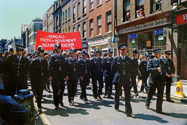 Protestors march with red banners behind a row of policemen.