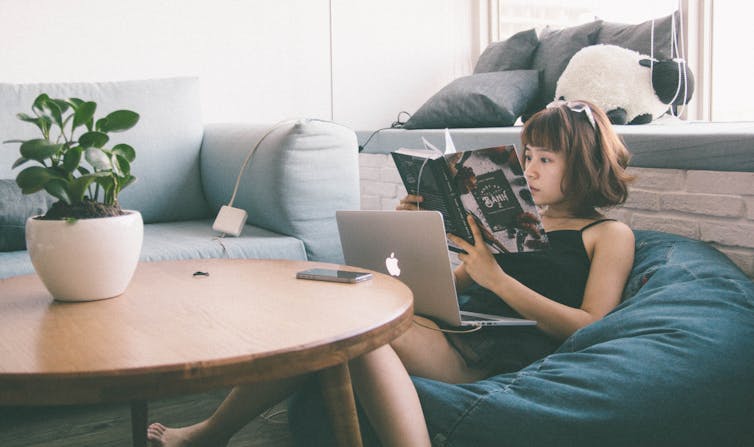 Woman sitting reading book, with laptop and phone.