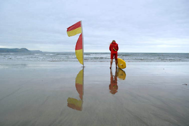 A person wearing red stands next to a yellow surfboard on a beach next to the edge of the sea. Two yellow and red flags fly on a pole next to them.