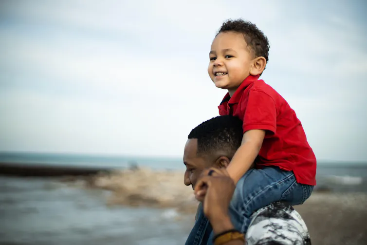 A young father with his mixed-heritage son on his shoulders, smiling.