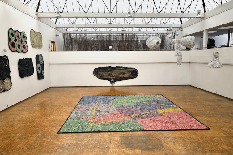 A gallery with a huge square work on the floor, tapestry-like sculptures on one wall and hanging sculptures opposite.