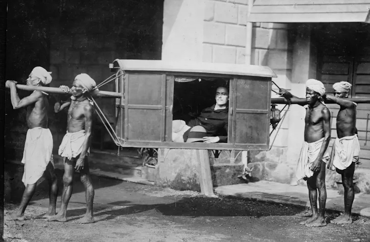 A group of Indian servants carrying a large white man in a sedan carriage.
