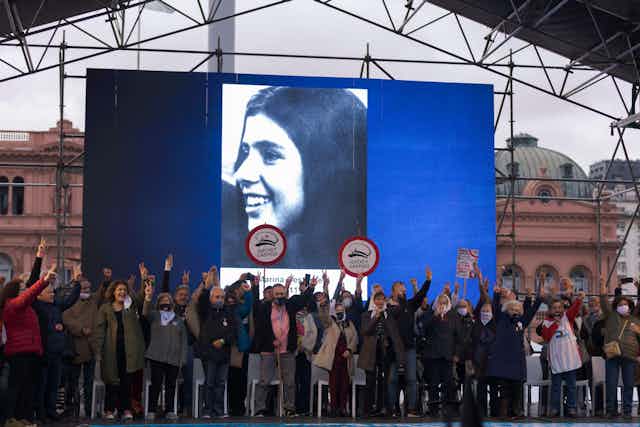 A group of people standing in a square holding their hands in the air, some holding banners, in front of a large poster for one of the people who was disappeared during Argentina's dictatorship.