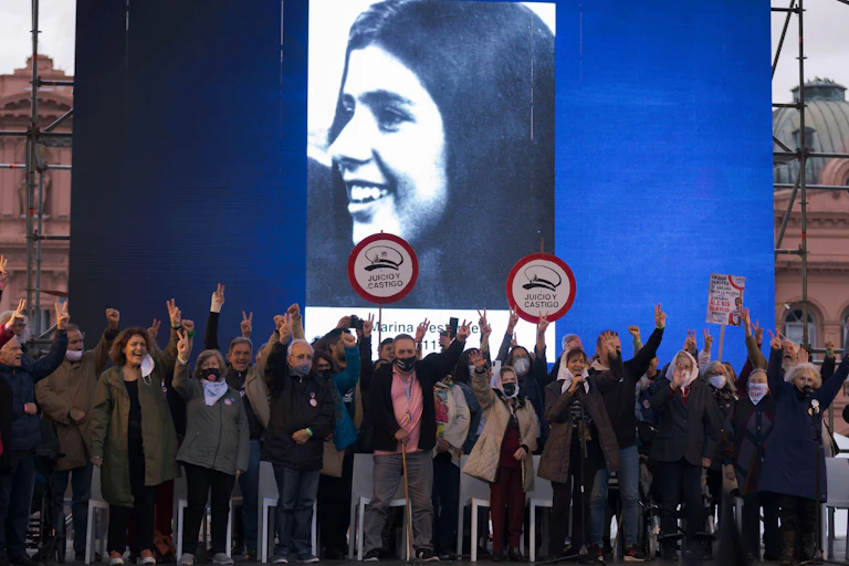 A group of people standing in a square holding their hands in the air, some holding banners, in front of a large poster for one of the people who was disappeared during Argentina's dictatorship.